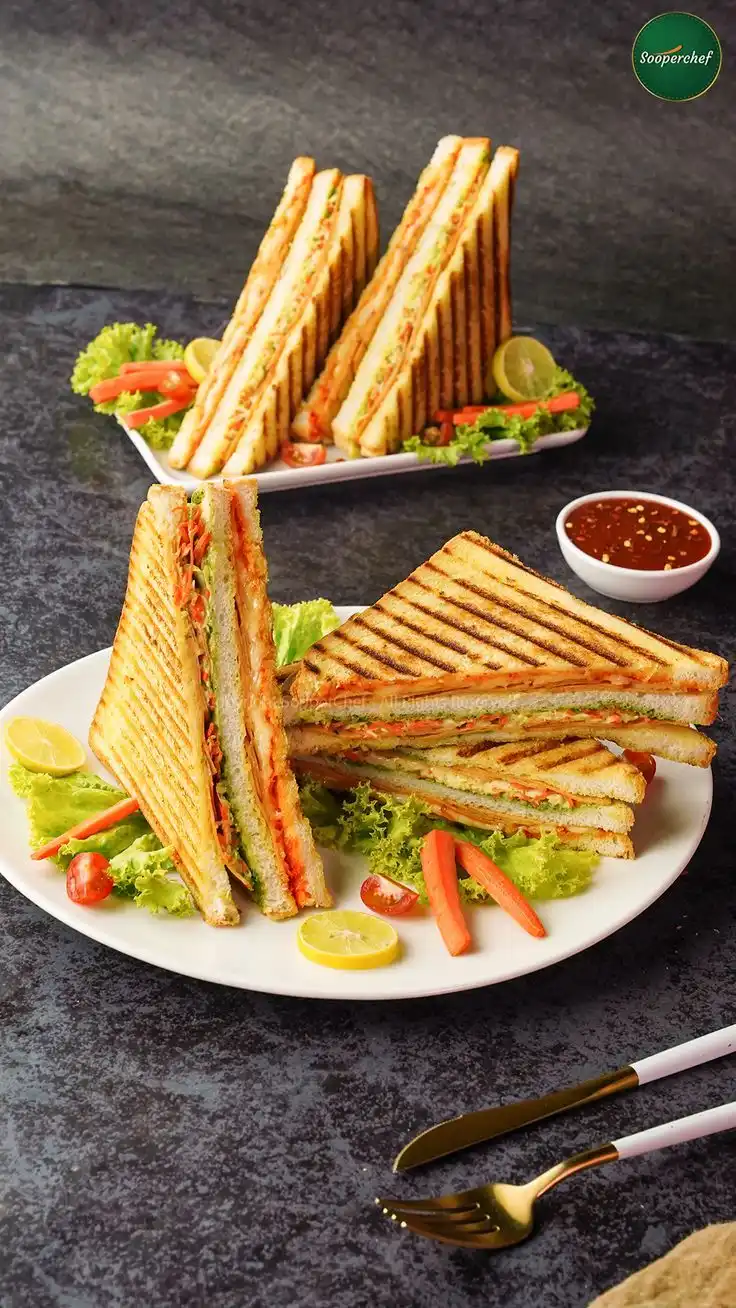 Grilled triple-decker sandwich served on a white plate with lettuce, cherry tomatoes, and carrot slices.